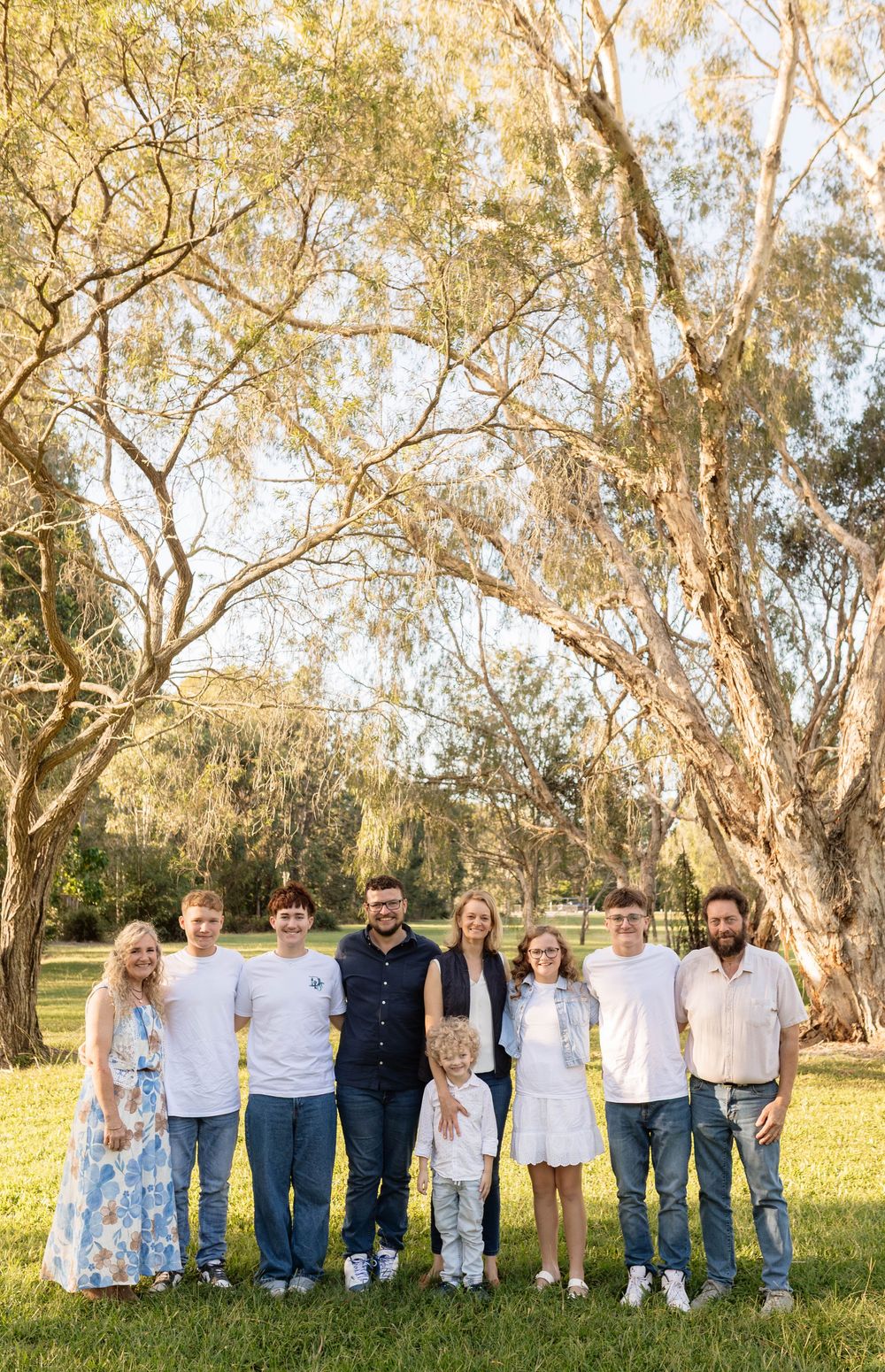 A joyful family of eight poses together outdoors in a sunny park surrounded by tall trees on green grass.