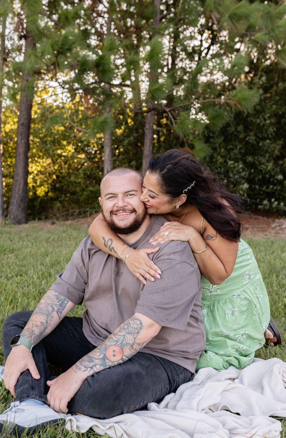 A tattooed couple shares a romantic moment on a blanket in a park, with the woman kissing the smiling man on the cheek.
