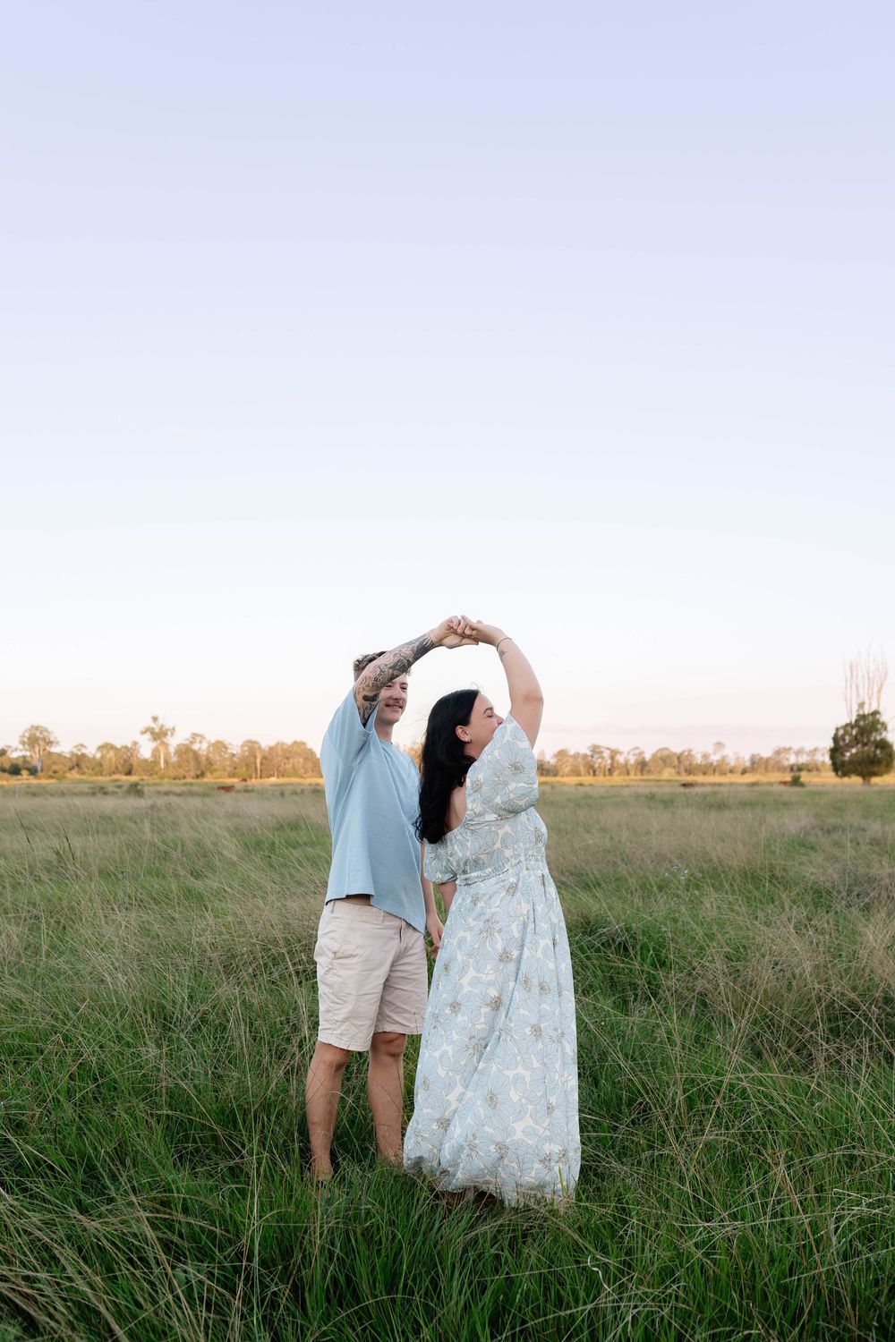 Couple dancing together in a green field at sunset, woman in floral dress and man in light blue shirt holding hands overhead.