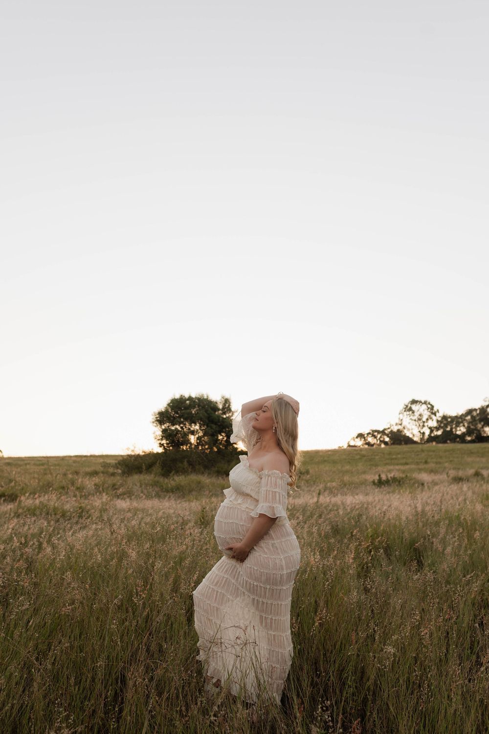 Pregnant woman in white lace off-shoulder gown standing in golden field at sunset, cradling baby bump.