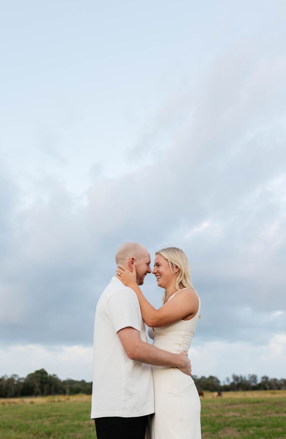 A couple embraces and smiles at each other in an open field under a cloudy sky, dressed in white outfits.