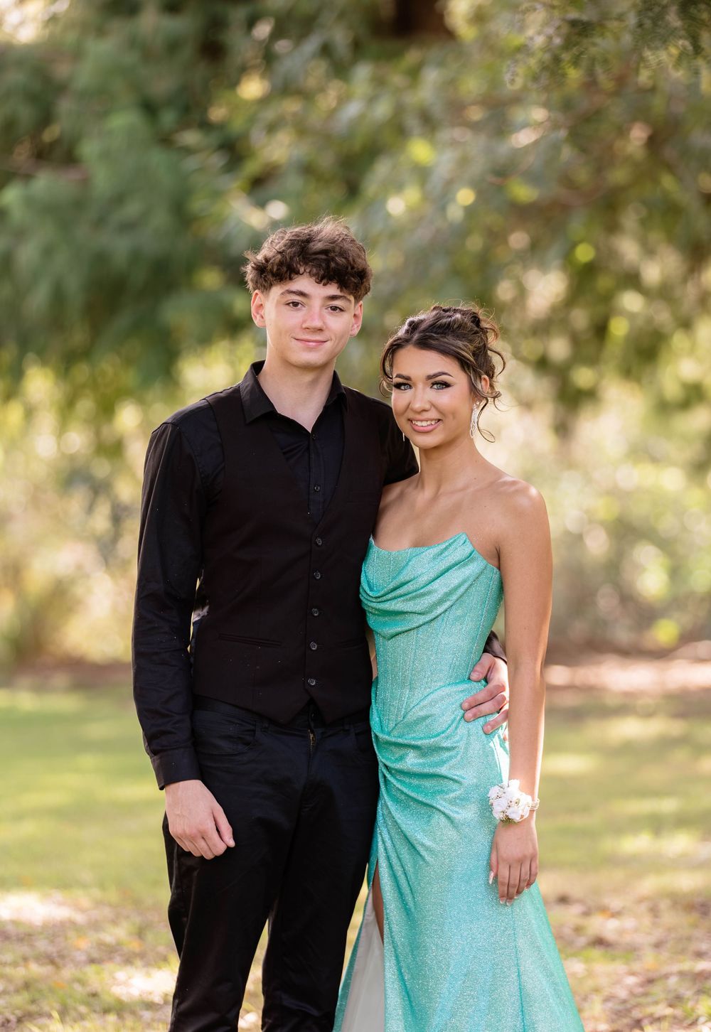 A teenage couple dressed in prom attire poses outdoors, him in black and her in a teal gown.