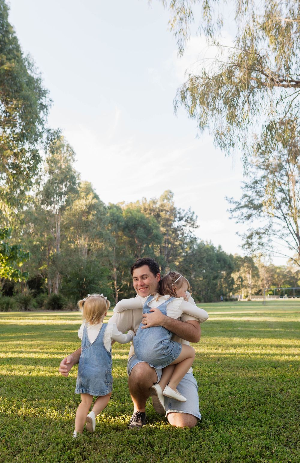A parent crouches on green grass hugging two young children in denim overalls outdoors in a sunny park.