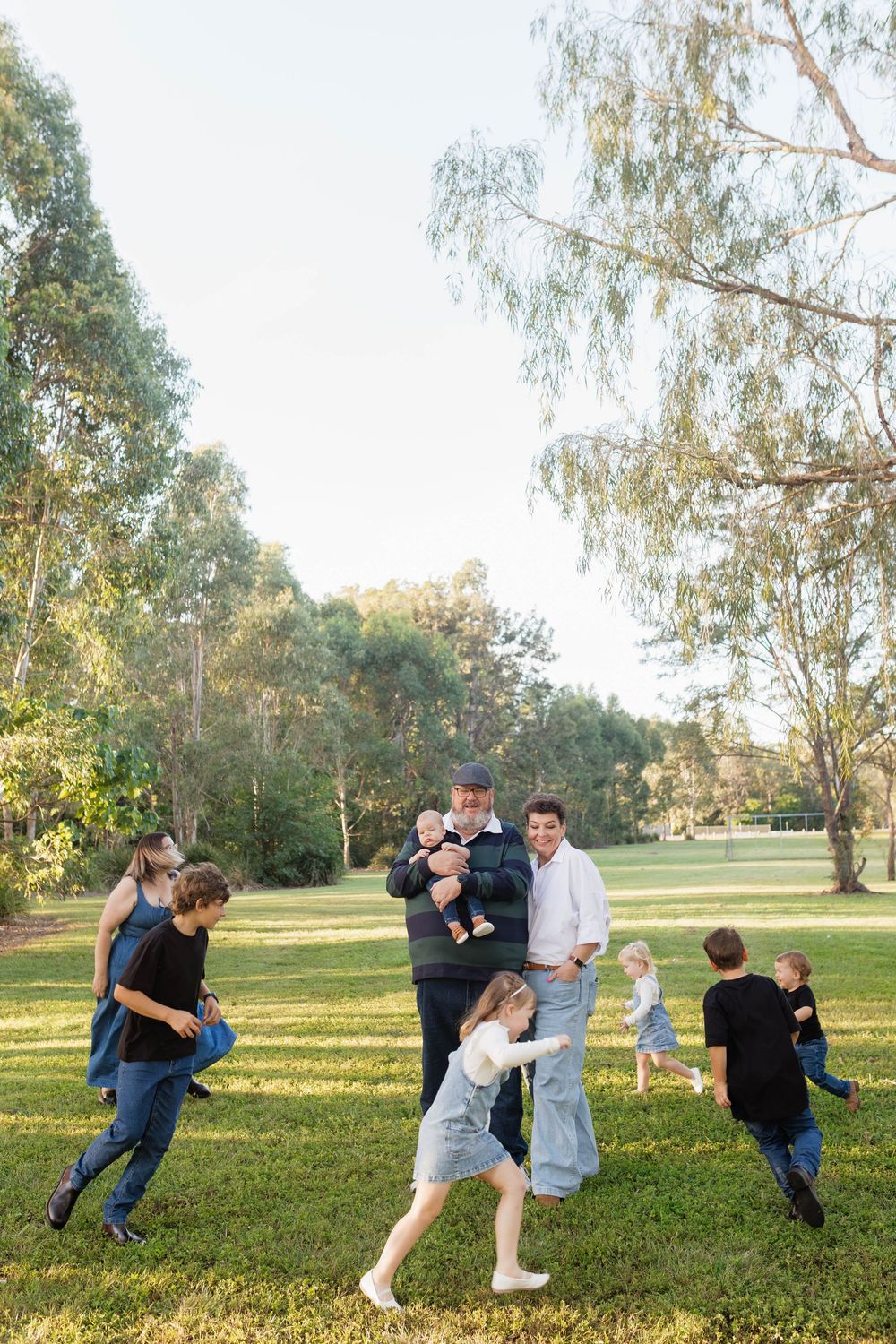 A joyful family plays together on a lush green lawn surrounded by tall trees during a sunny outdoor photo session.
