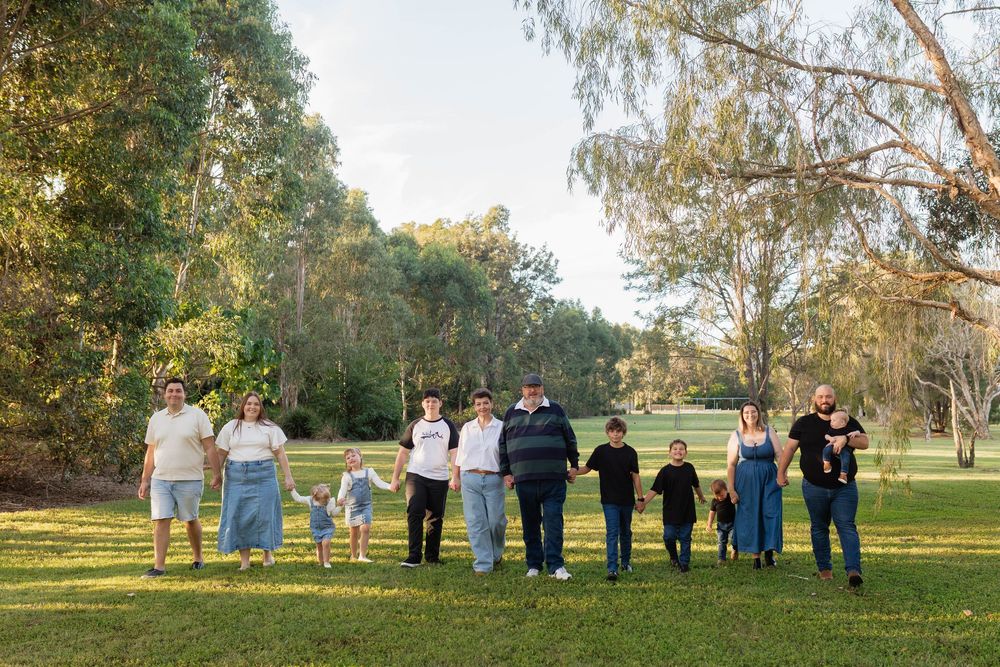 Large family group holding hands in a line outdoors in a lush green park surrounded by tall trees.