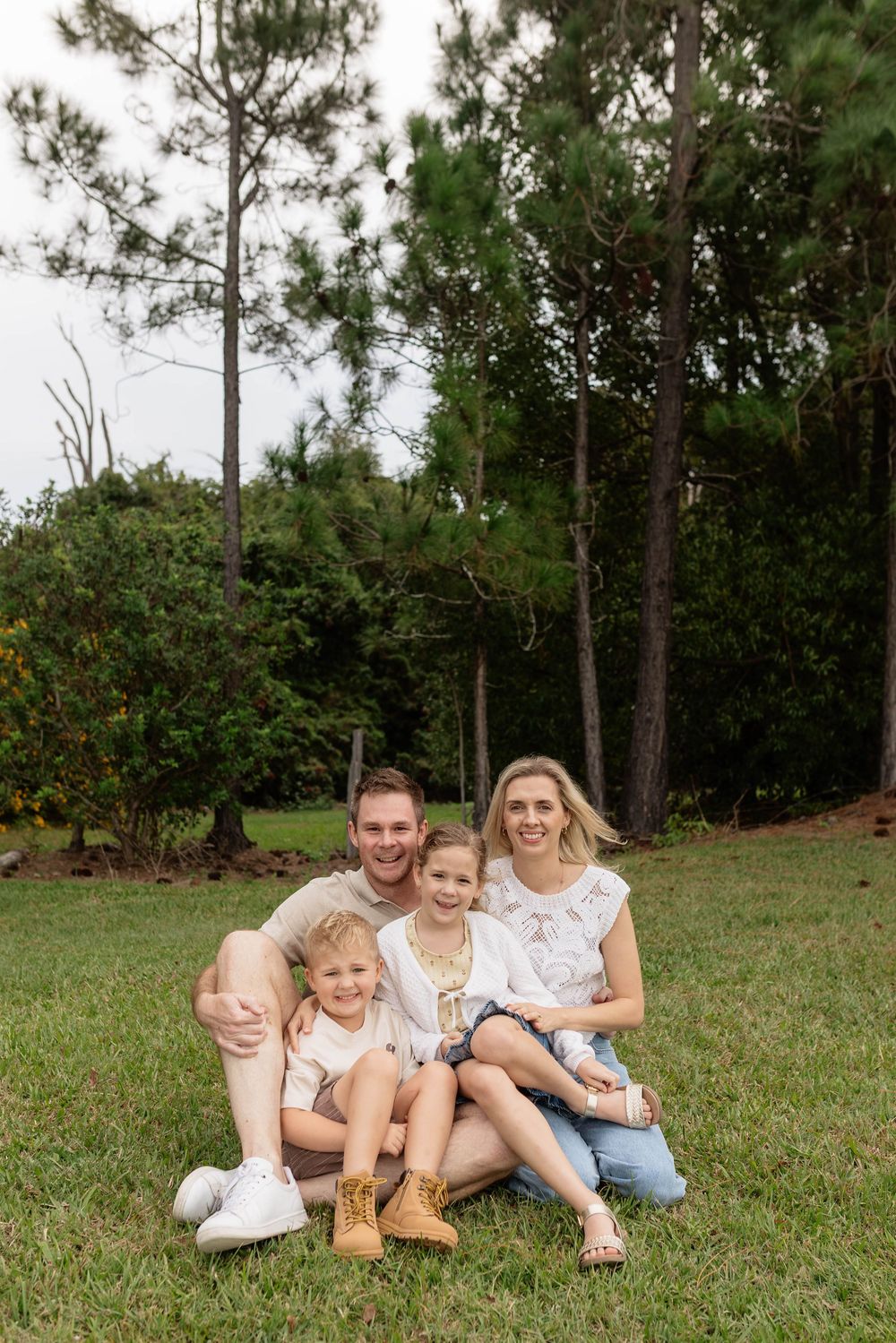 Happy family of four sitting together on green grass outdoors, smiling in front of tall pine trees.