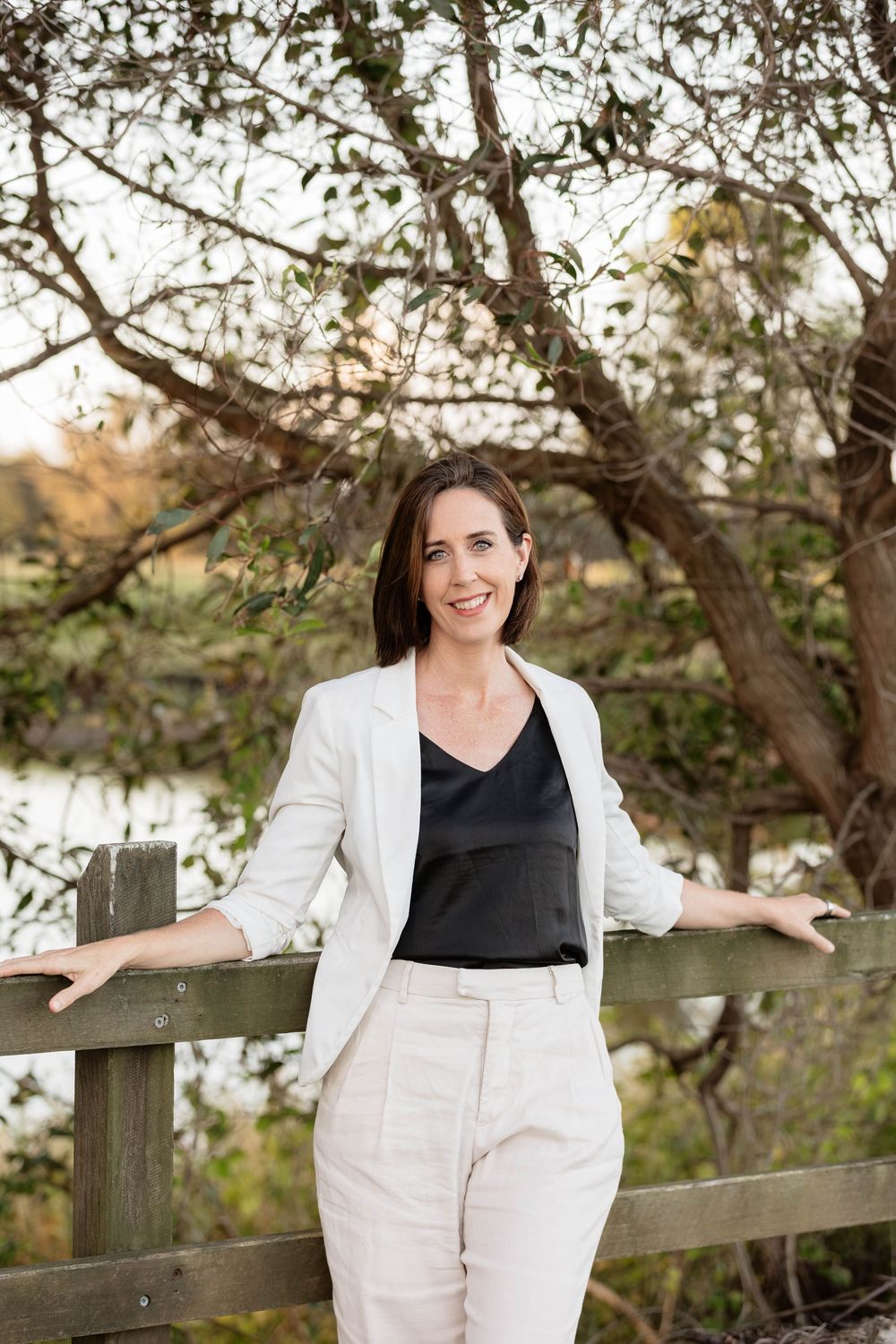 Woman in white blazer and pants smiling outdoors by wooden fence with trees in background.