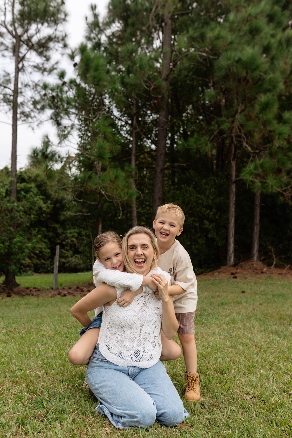 A joyful mother kneels on grass while her two young children hug her from behind, laughing outdoors near tall pine trees.