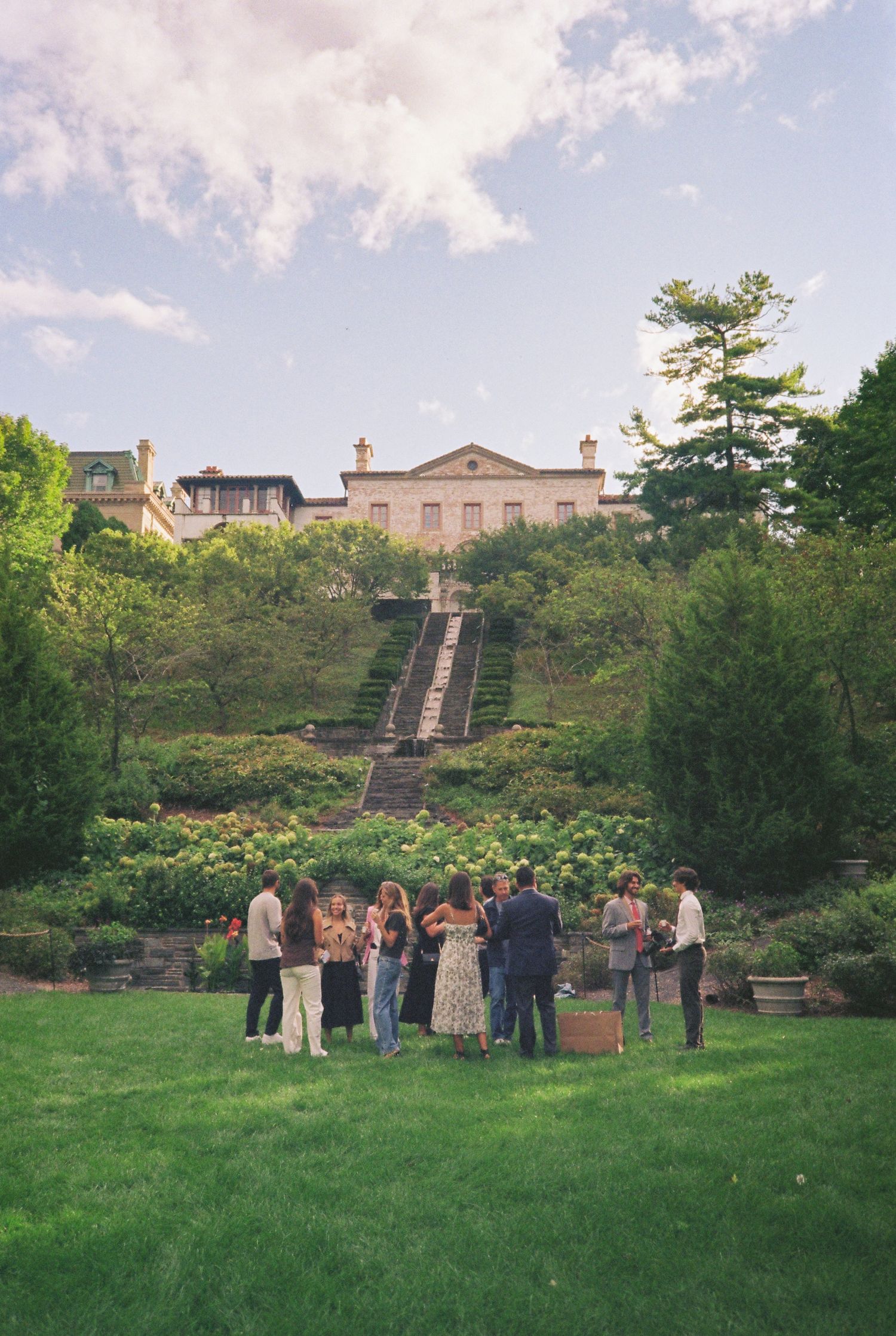 Guests mingle on lush green lawn below grand estate mansion with stone staircase and terraced gardens at outdoor event.