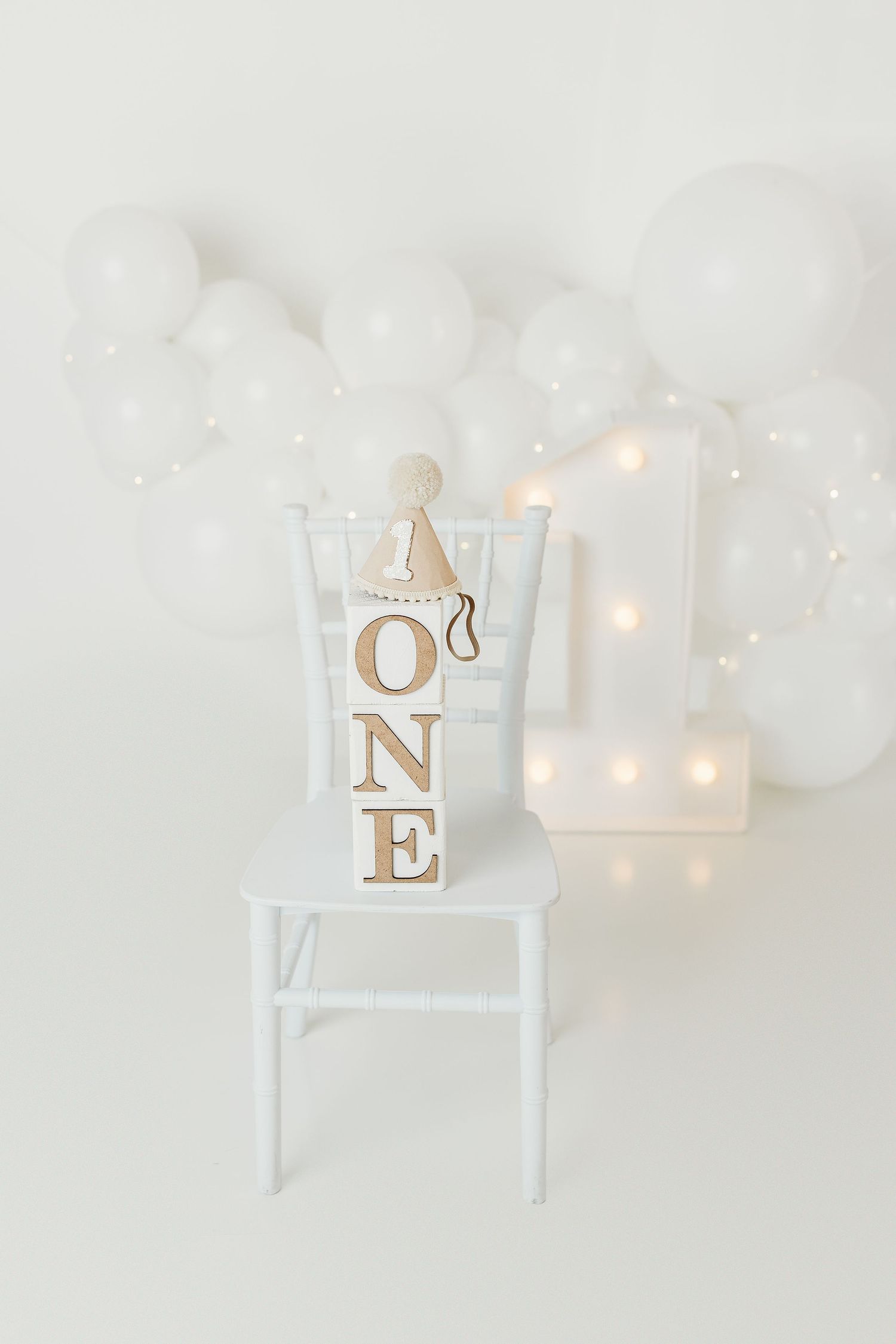 White first birthday setup with ONE letter blocks on a chair, balloon garland, and glowing number one light backdrop.