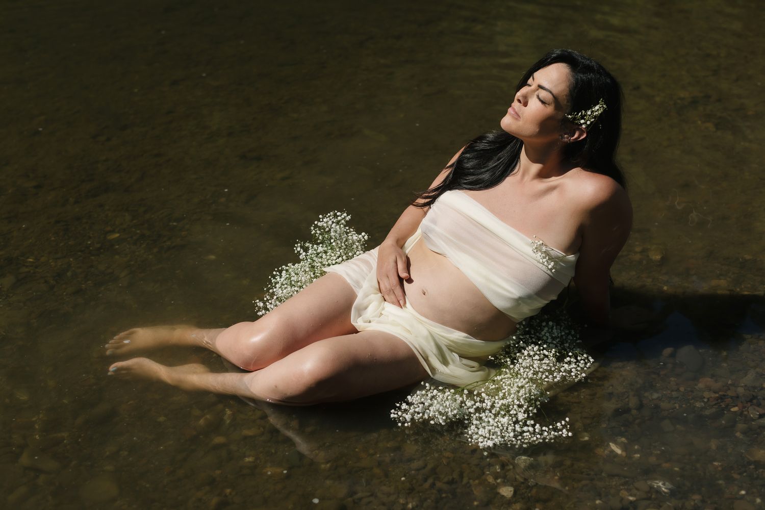 Expectant mother wears cream mesh covering for maternity photos laying in a shallow river. North Conway Lakes Region maternity photographer.