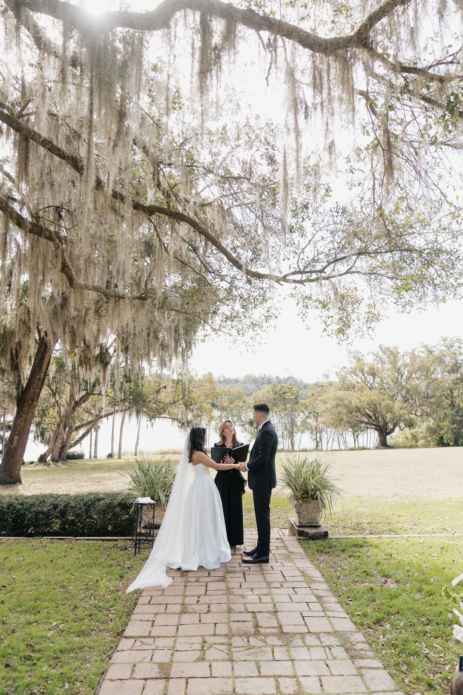 Bride and groom exchange vows under Spanish moss-draped oak trees on a brick path at an outdoor wedding ceremony.