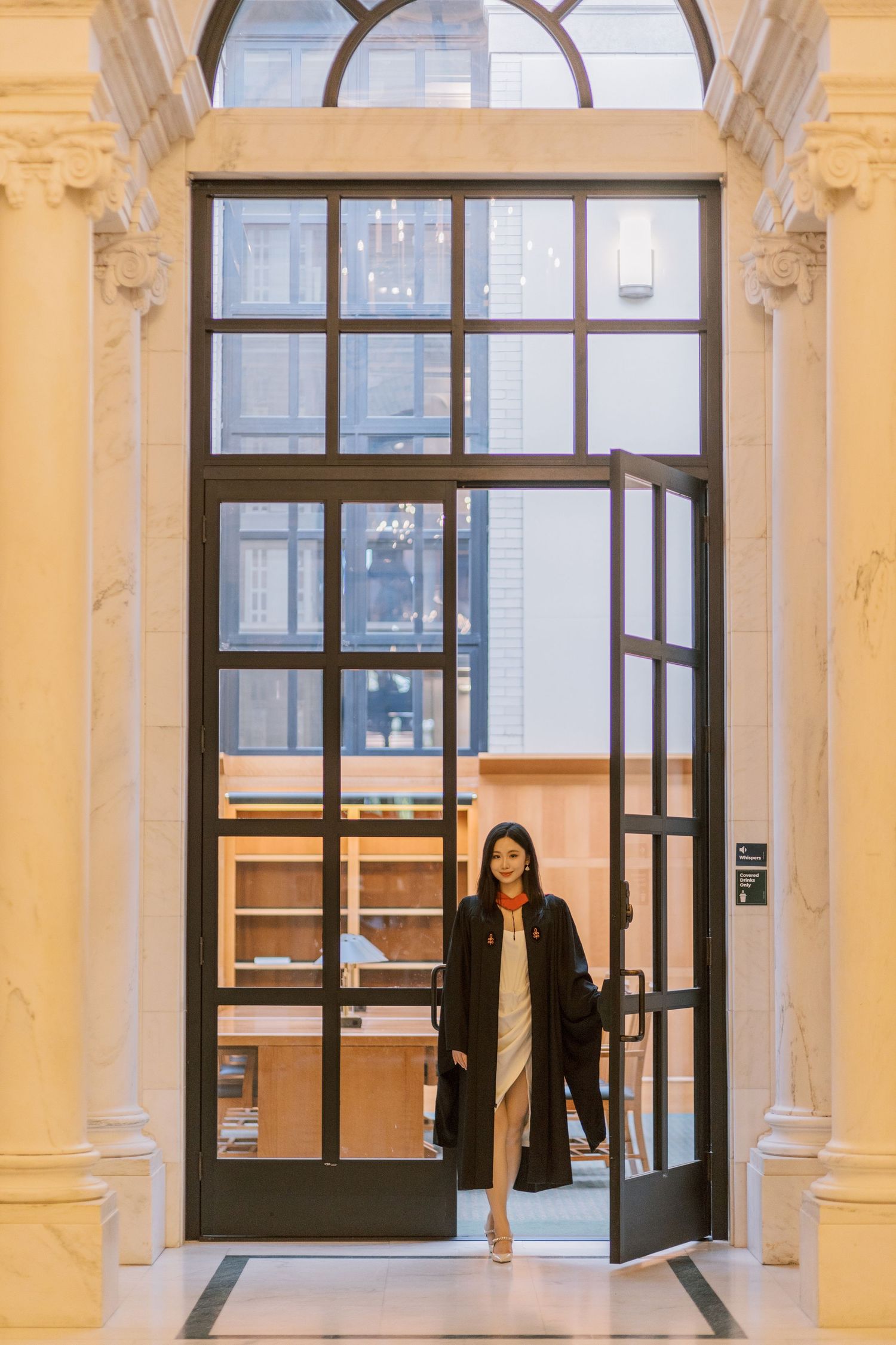 Elegant woman in a Harvard graduation robe standing in grand doorway with floor-to-ceiling windows and marble columns.
