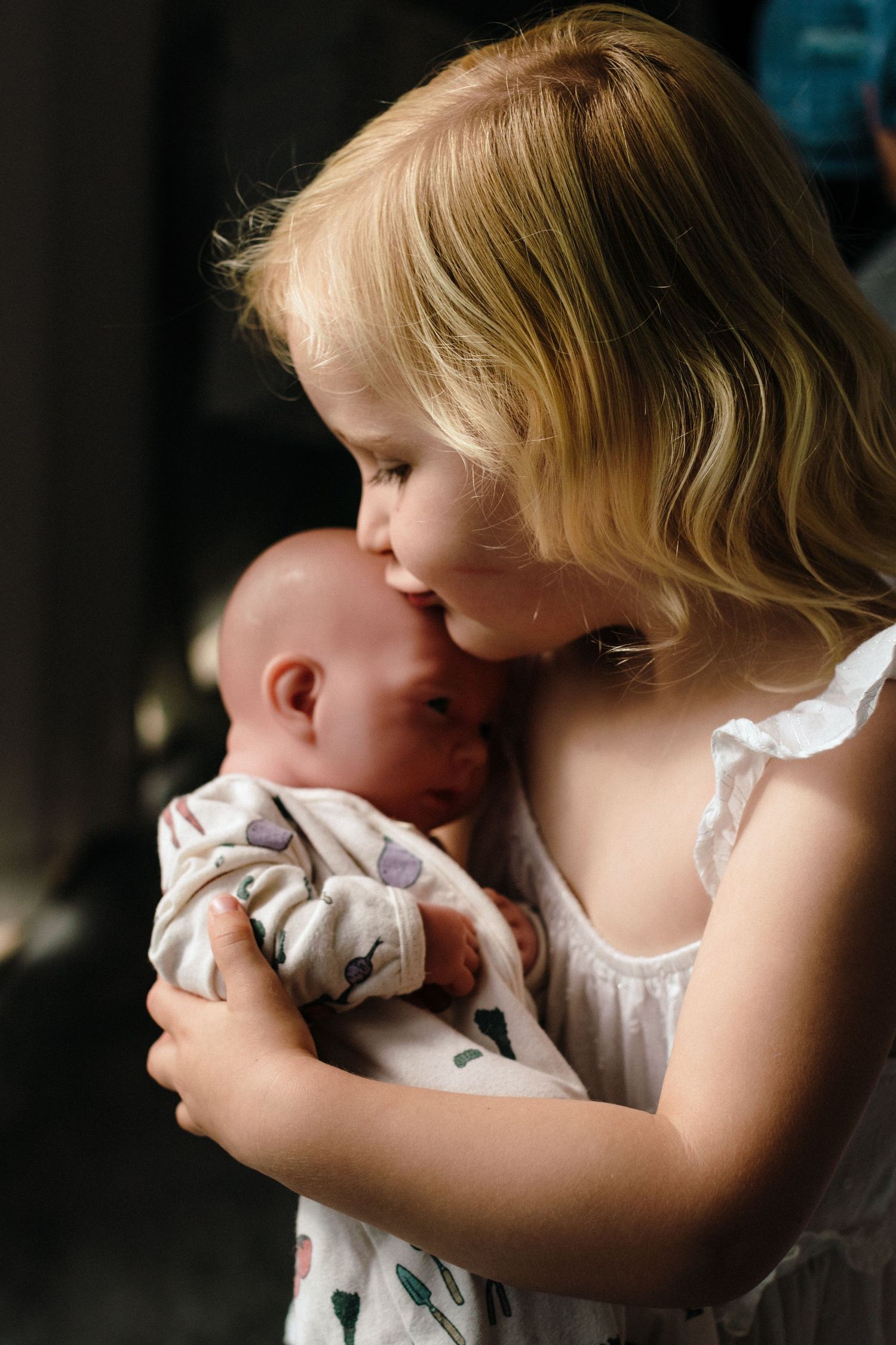 Young blonde girl in white dress gently kisses newborn baby wrapped in floral swaddle blanket.