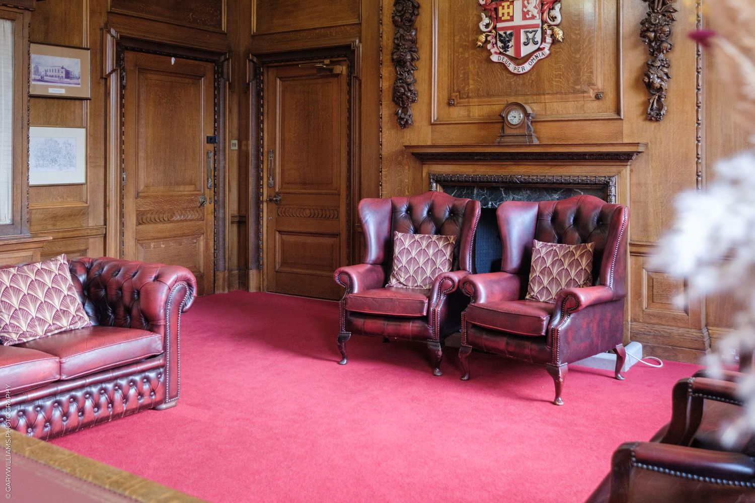 Elegant room with red Chesterfield sofas and armchairs on pink carpet beside a wooden fireplace mantel.