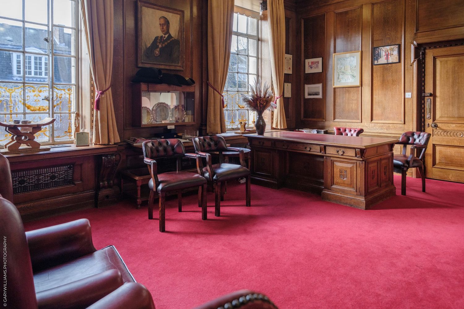 Elegant traditional office with red carpet, leather chairs, wooden desk, and portrait painting in a grand historic building.