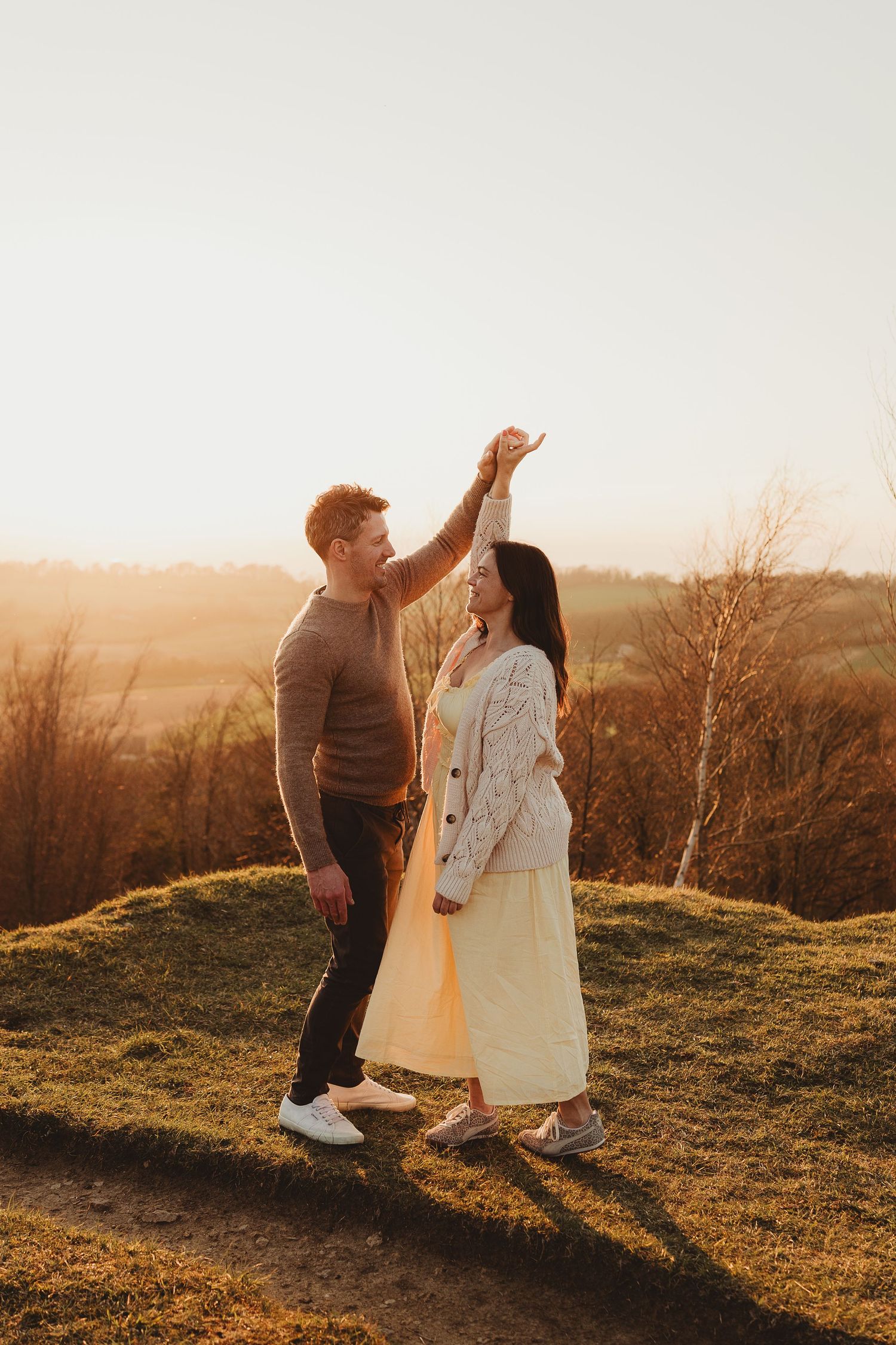 Couple dancing at golden hour on a grassy hill, man spinning woman in yellow skirt with autumn landscape behind.