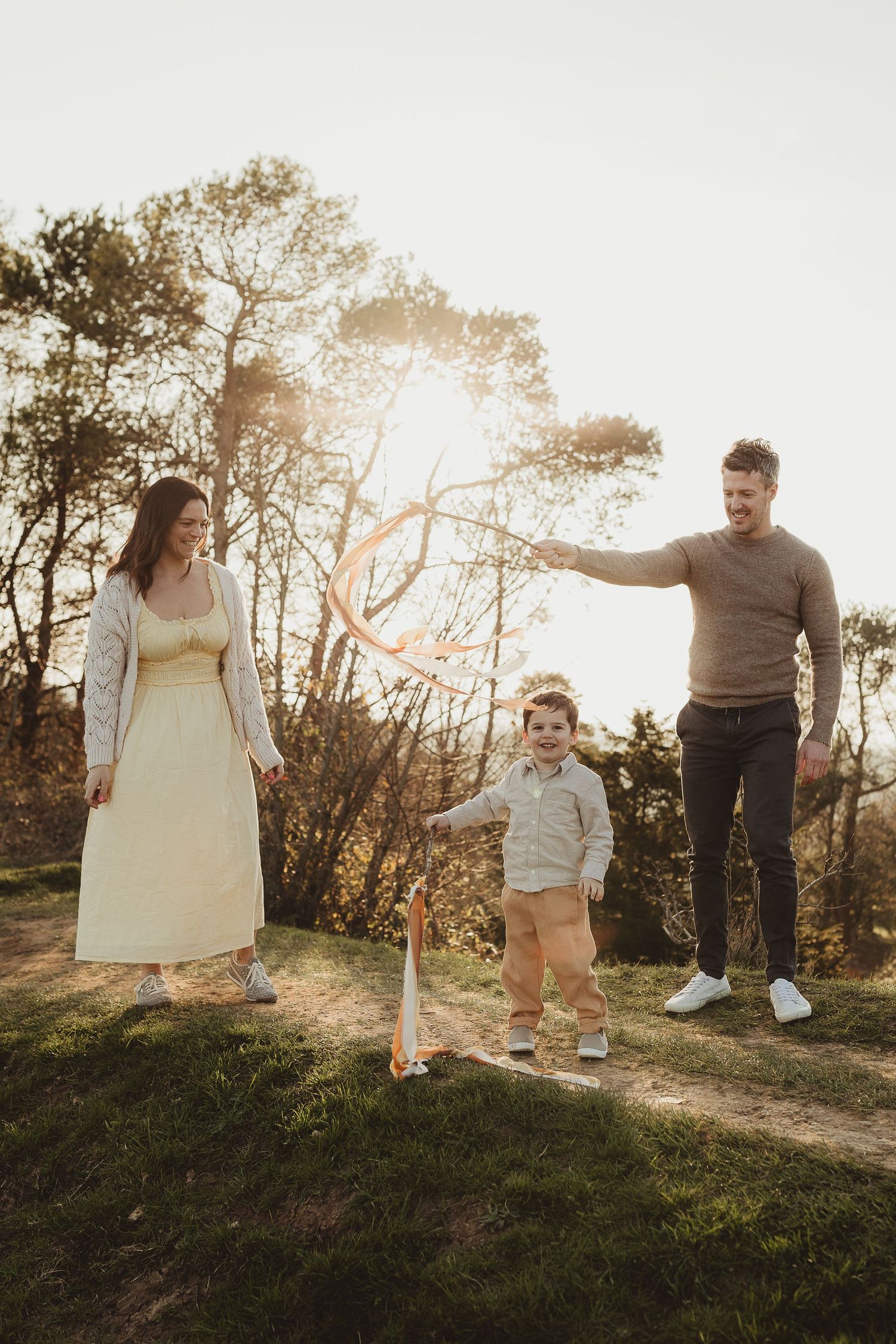 Family of three playing outdoors at golden hour, parents watching toddler with hula hoop among trees.