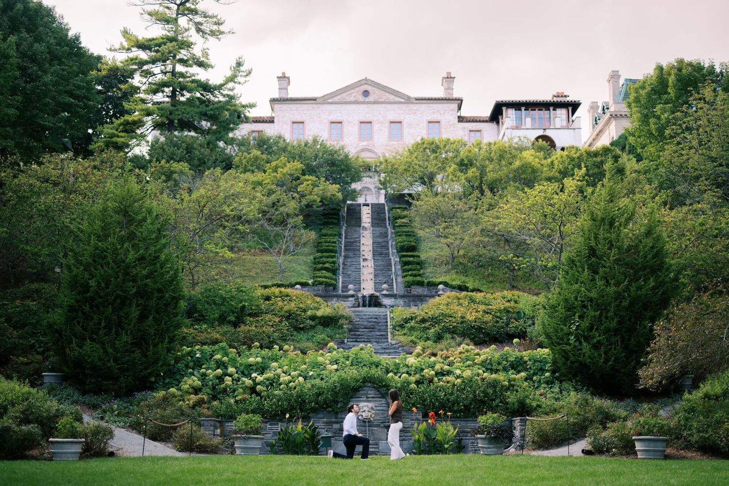 Couple poses in lush formal garden with grand staircase and elegant estate mansion in background.