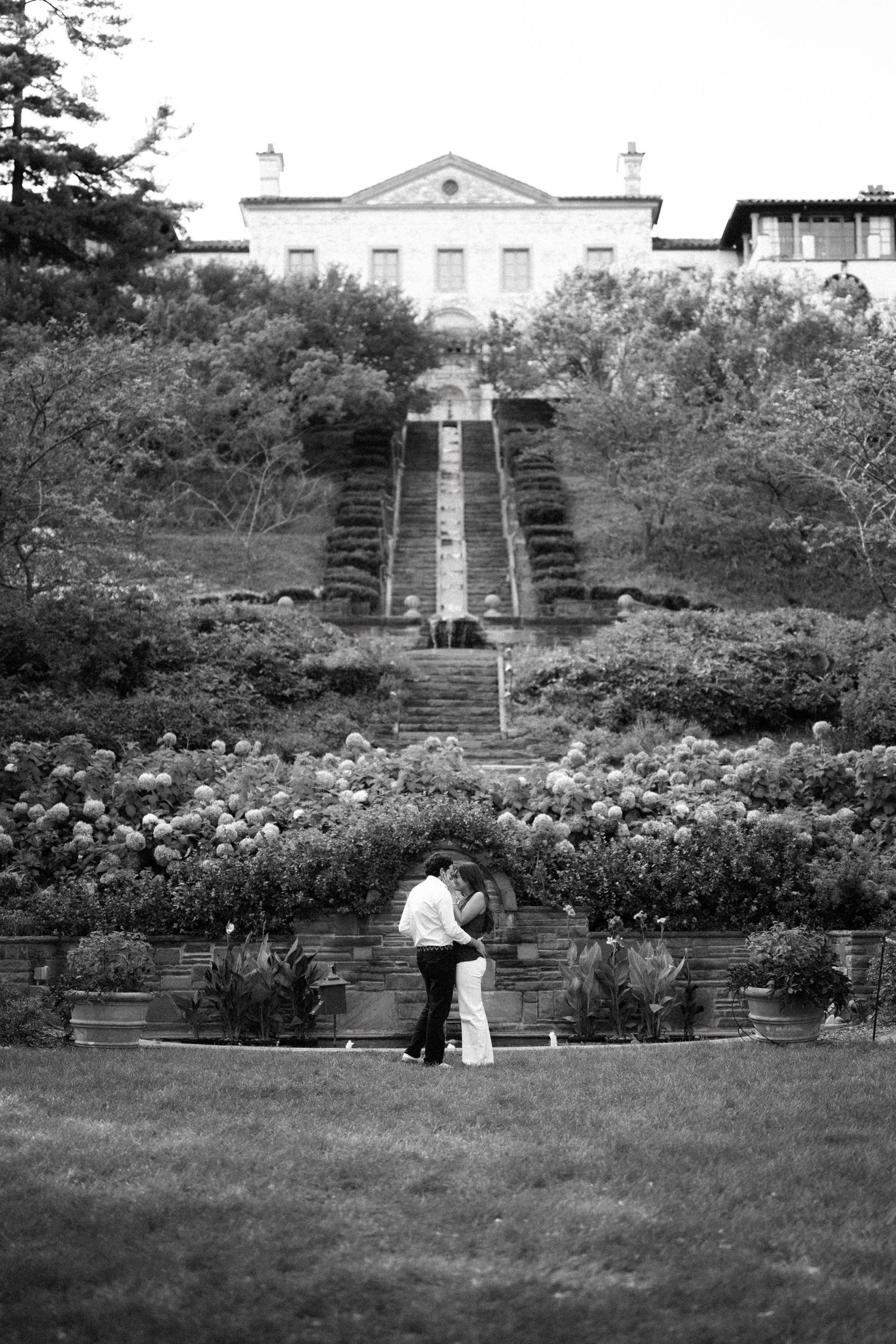 Black and white photo of a couple embracing in a formal garden with grand staircase and elegant estate in background.
