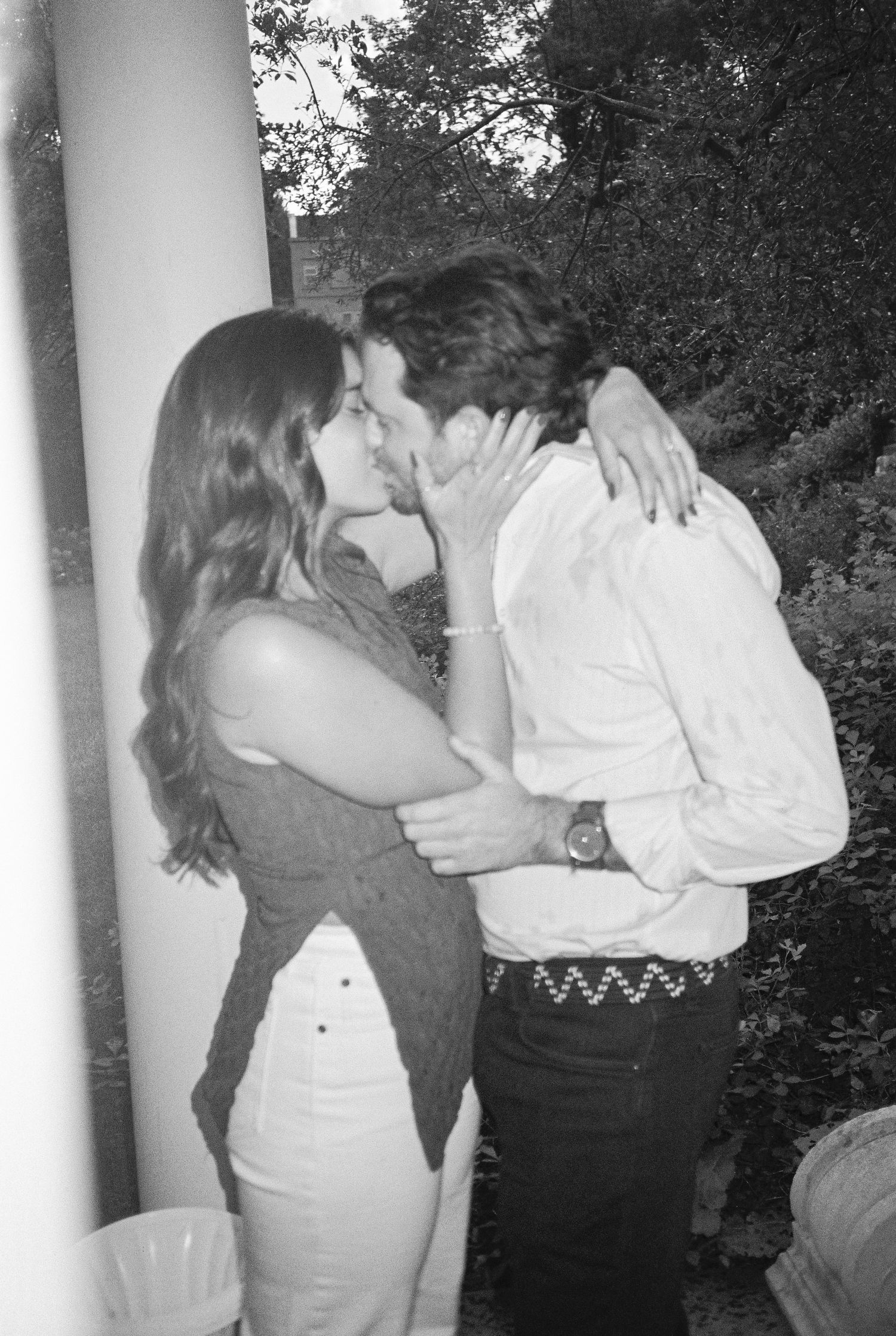 Black and white photo of a couple sharing a romantic kiss outdoors at a social gathering.