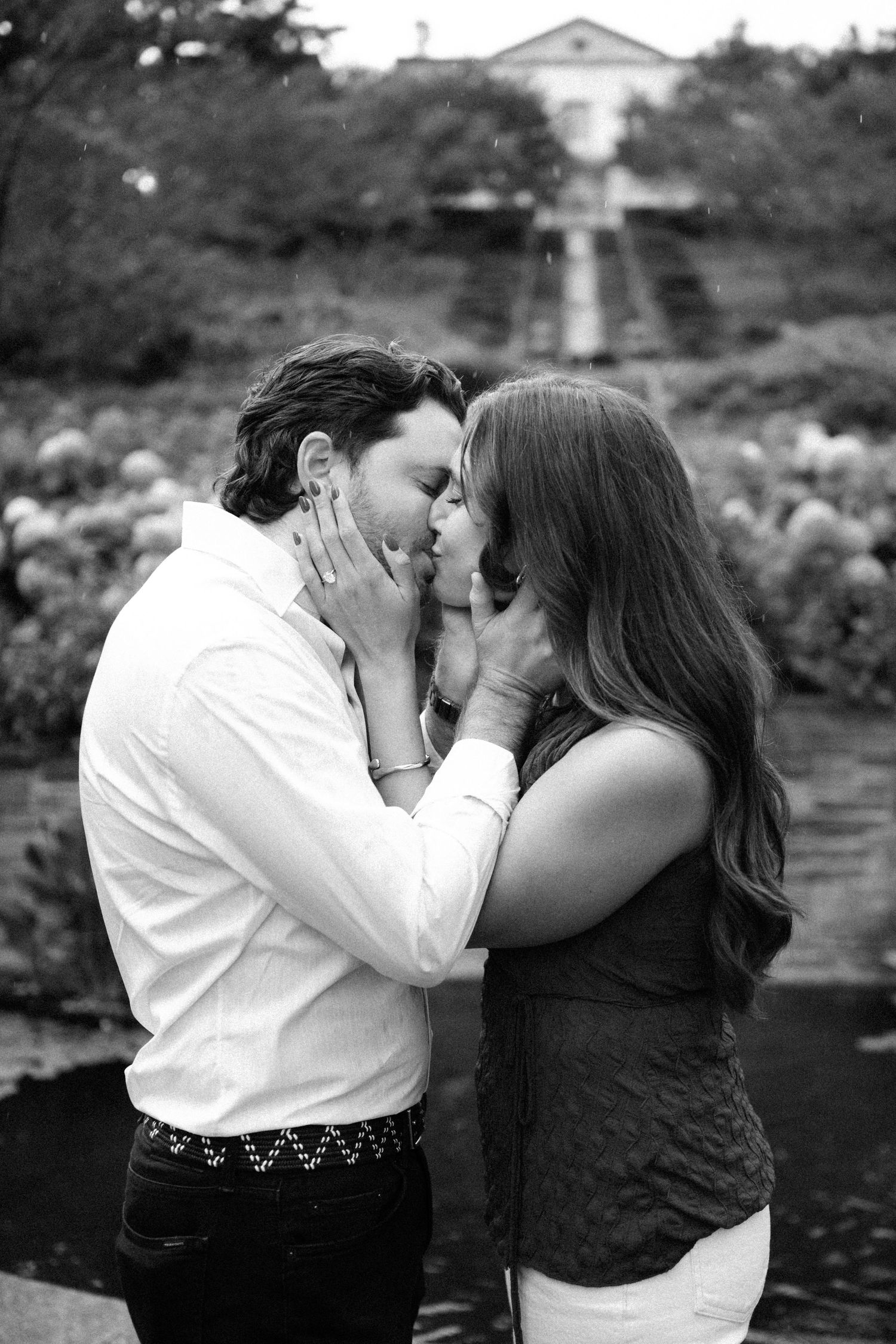 Black and white photo of a couple sharing a romantic kiss in a garden with a grand estate in the background.