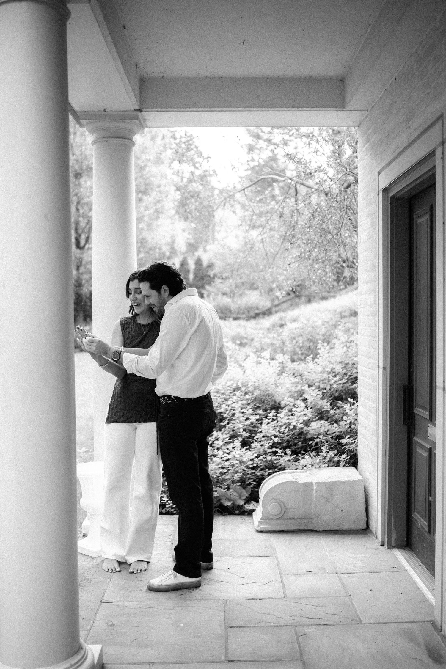 Black and white photo of a couple embracing on a classic columned porch surrounded by lush garden greenery.
