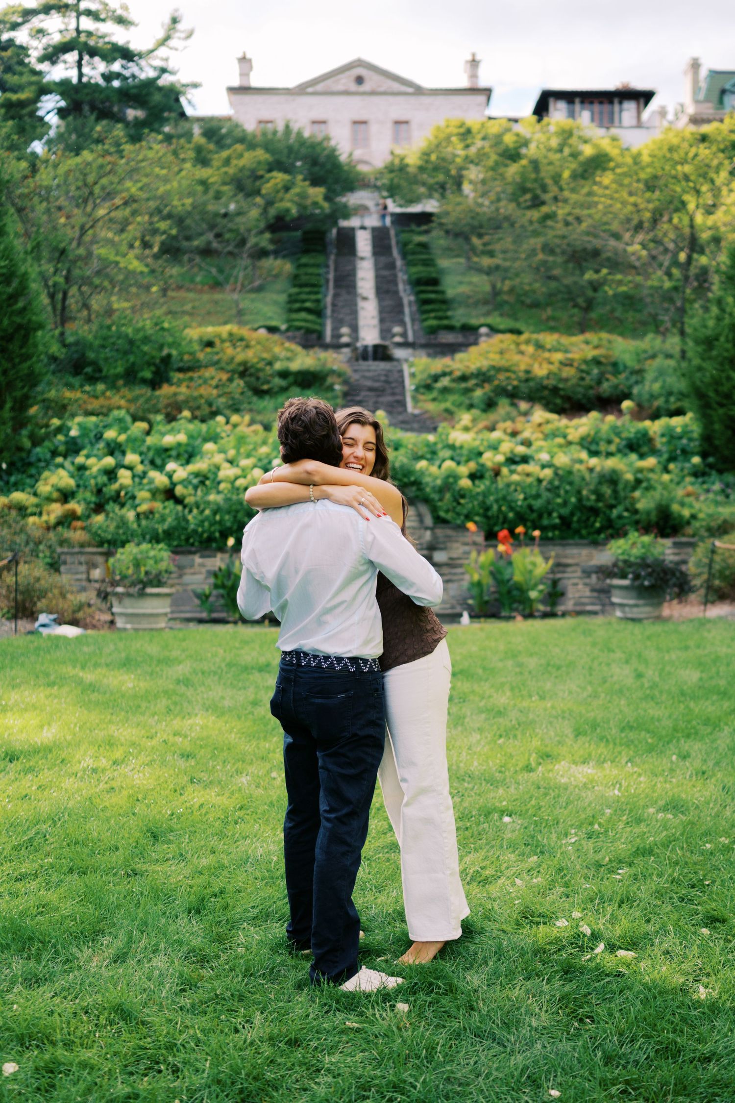 A couple embraces on a green lawn in front of a terraced garden with stone steps leading to an elegant estate.