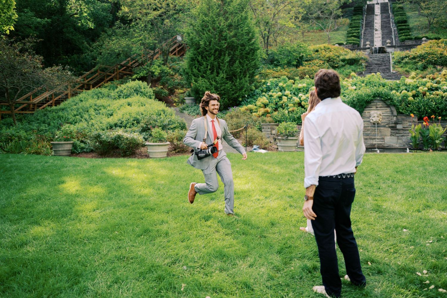 Two people playing in a lush green garden with terraced steps and colorful flowers in the background.