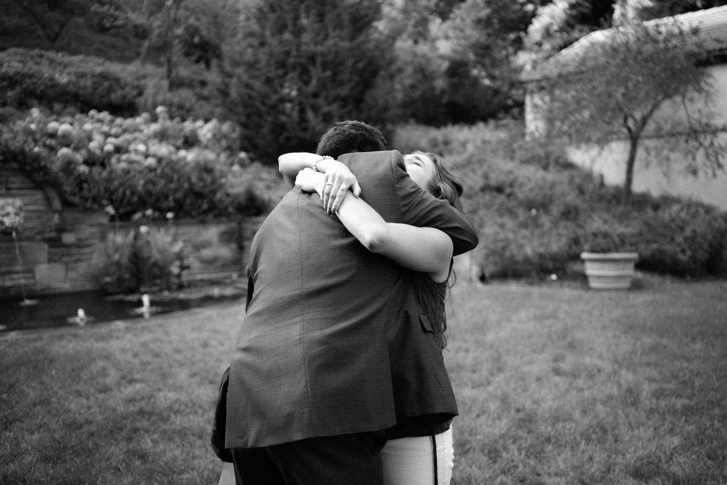 Two people share an emotional embrace in a garden setting, captured in black and white photography.
