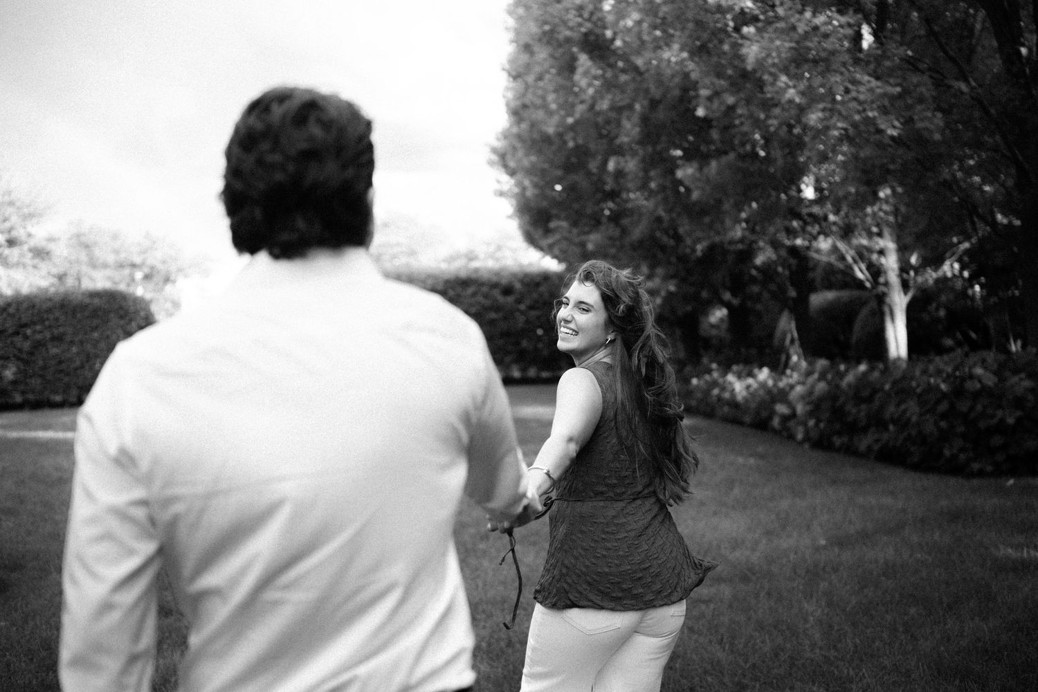 Black and white photo of a couple holding hands outdoors, woman looking back smiling at man in white shirt.