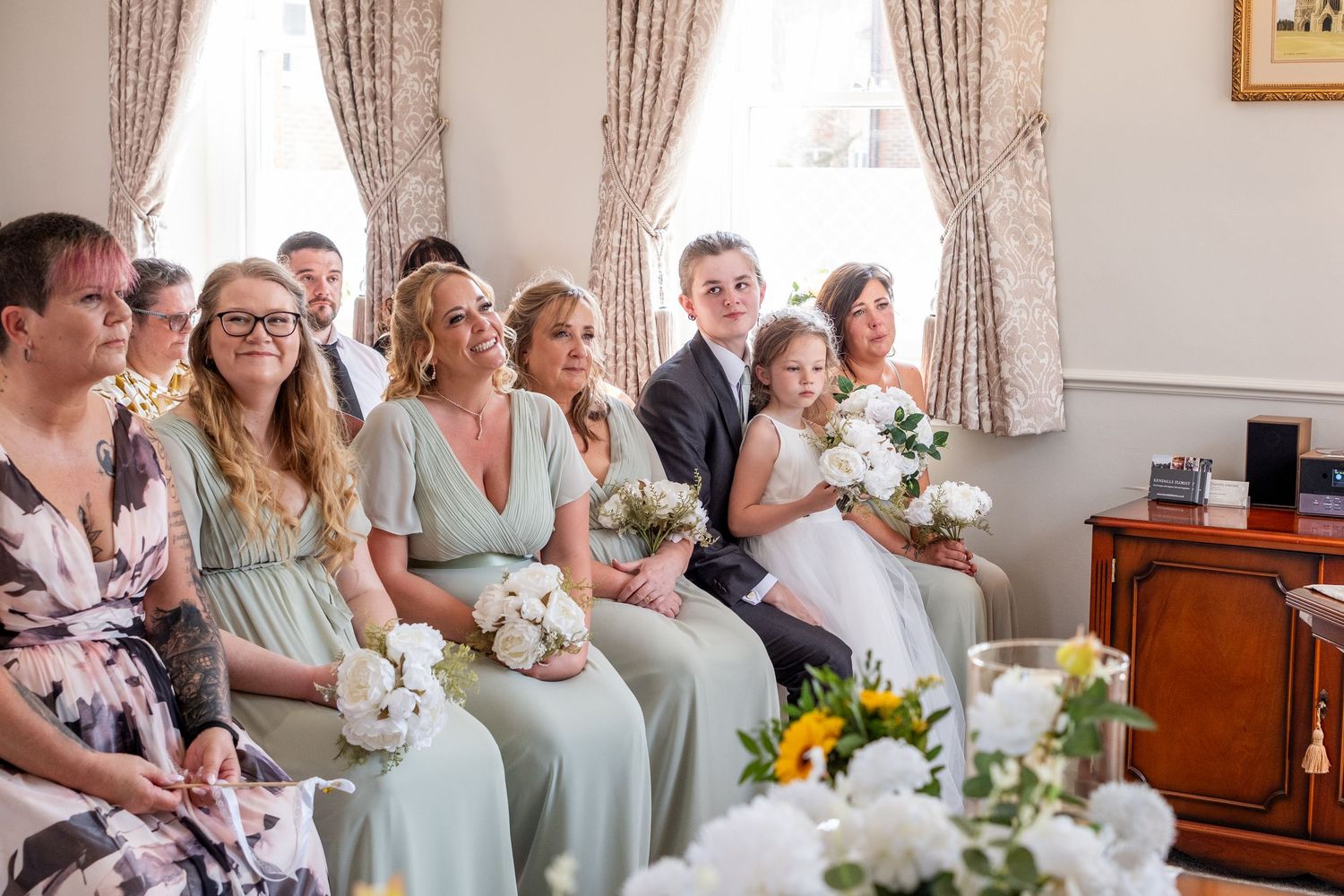 Wedding party seated indoors, bridesmaids in sage green dresses holding white bouquets, with guests smiling during a ceremony.