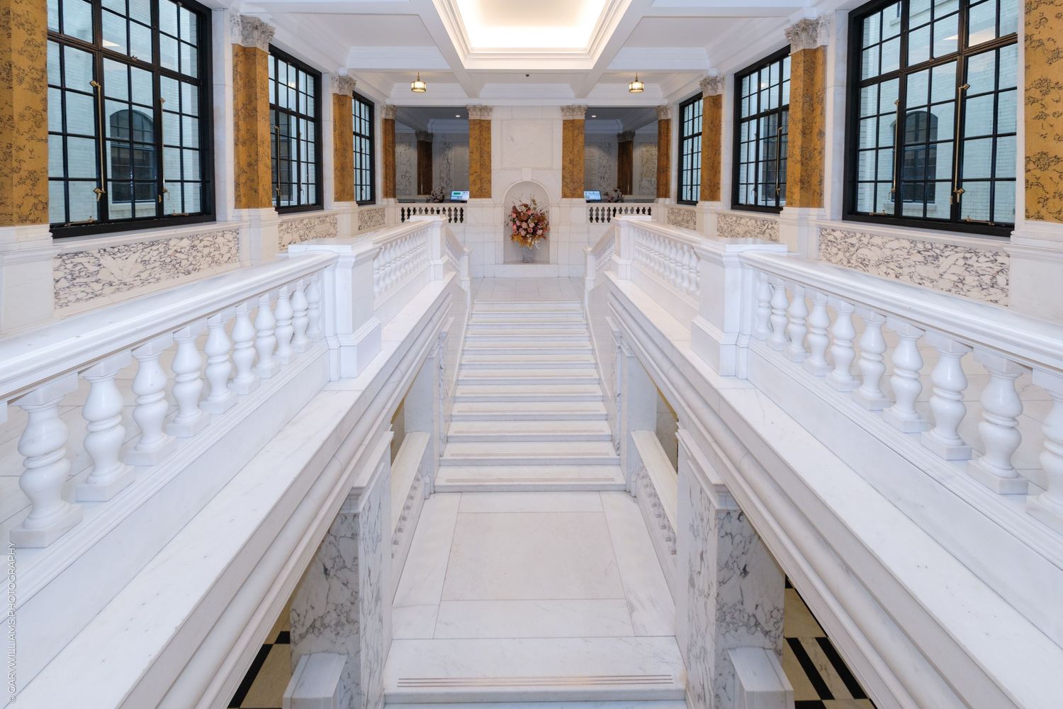 Grand white marble staircase with ornate railings in a classical building interior with tall windows.