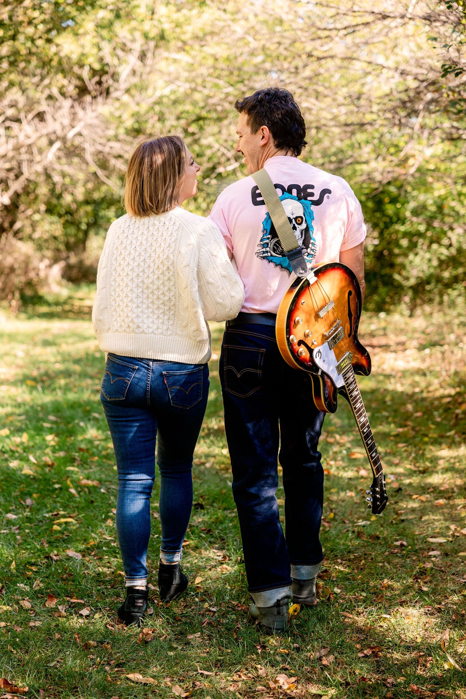 A couple walks together on an autumn path, with one carrying a guitar while wearing a pink t-shirt and jeans and the other in a sweater