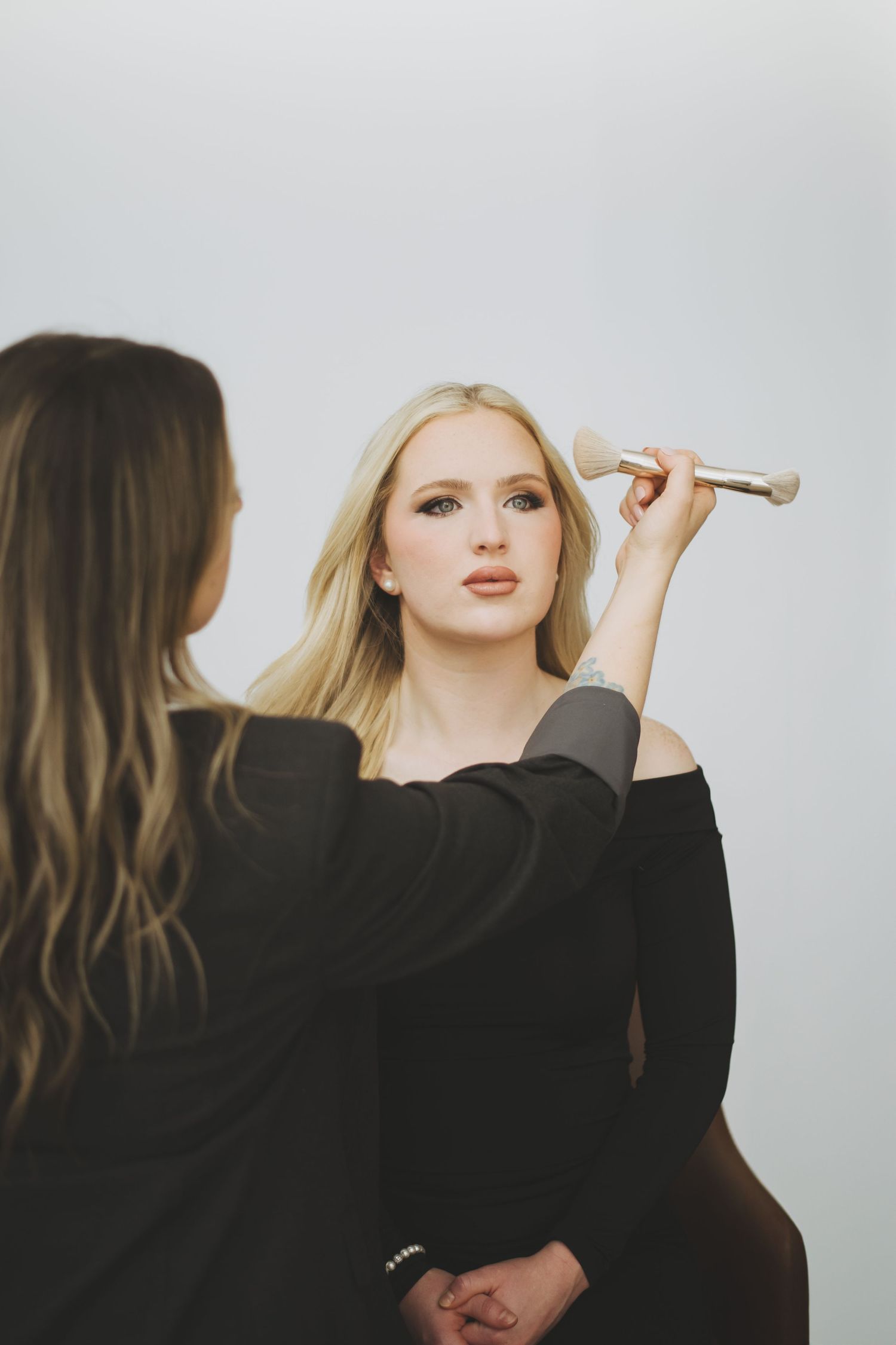 Makeup artist in black outfit applies cosmetics with brush against light background during professional beauty session.