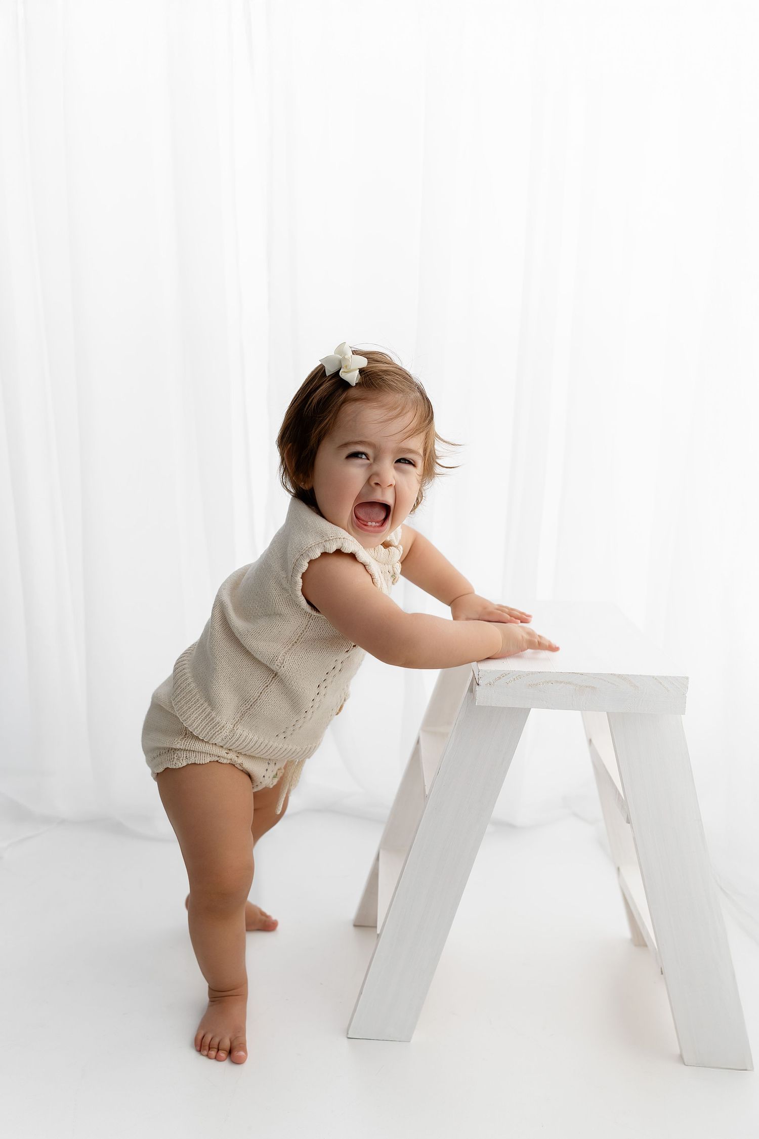 A toddler in a cream romper stands playfully beside a white chair on a white backdrop.