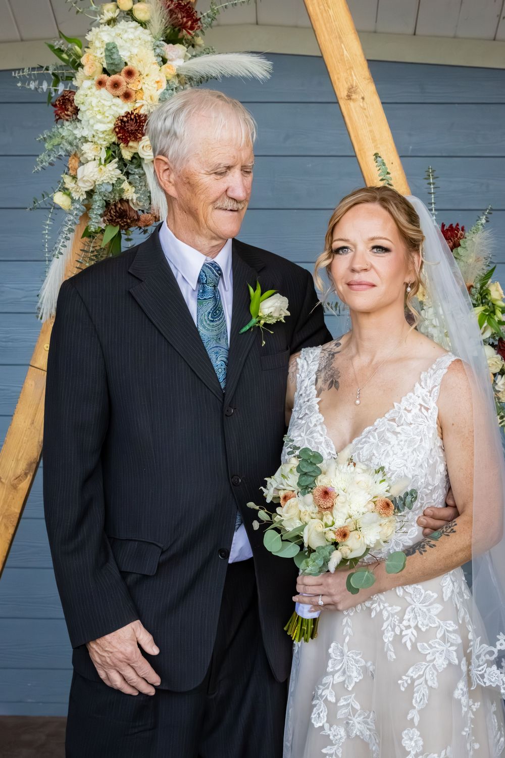 a bride looks at the camera, while her father in a black suit looks at her, in front of a wooden arch with flowers.