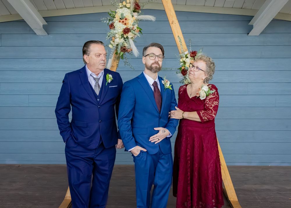 a groom standing in front of a wooden arch with flowers, smiling with his mother in a burgundy lace dress and his father in a navy blue suit
