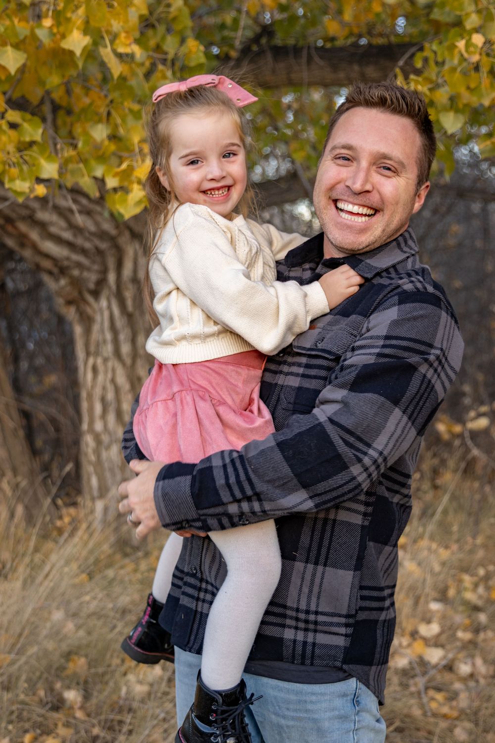 a young girl giggles while being held by her father, standing in front of a autumn tree.