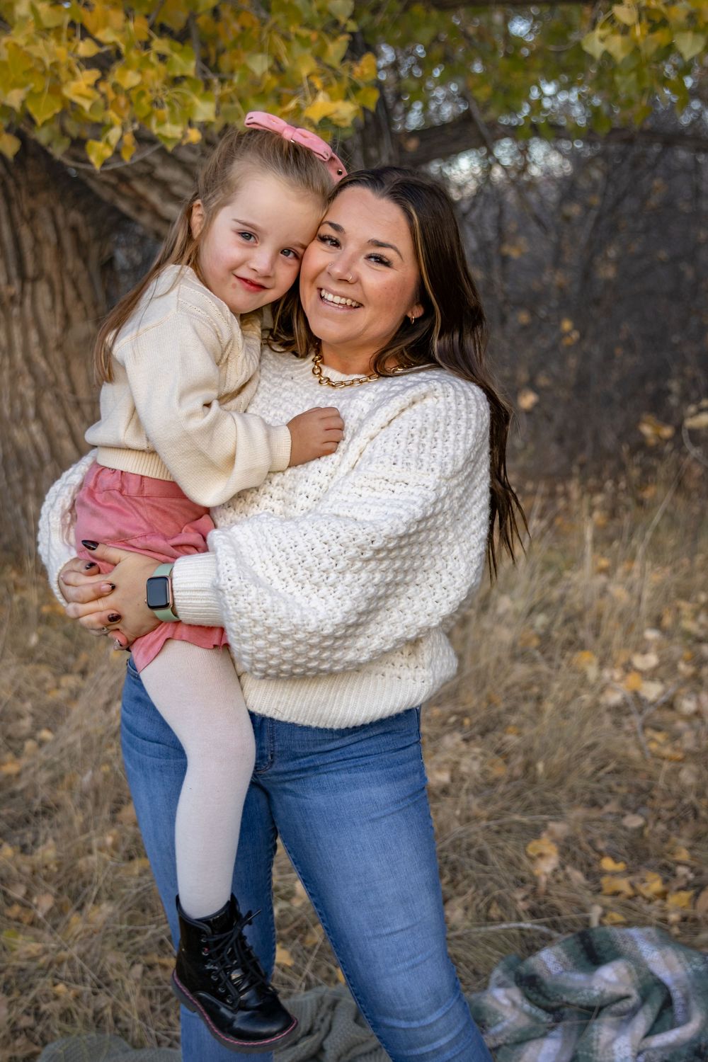 a young girl giggles while being held by her mother, standing in front of a autumn tree.
