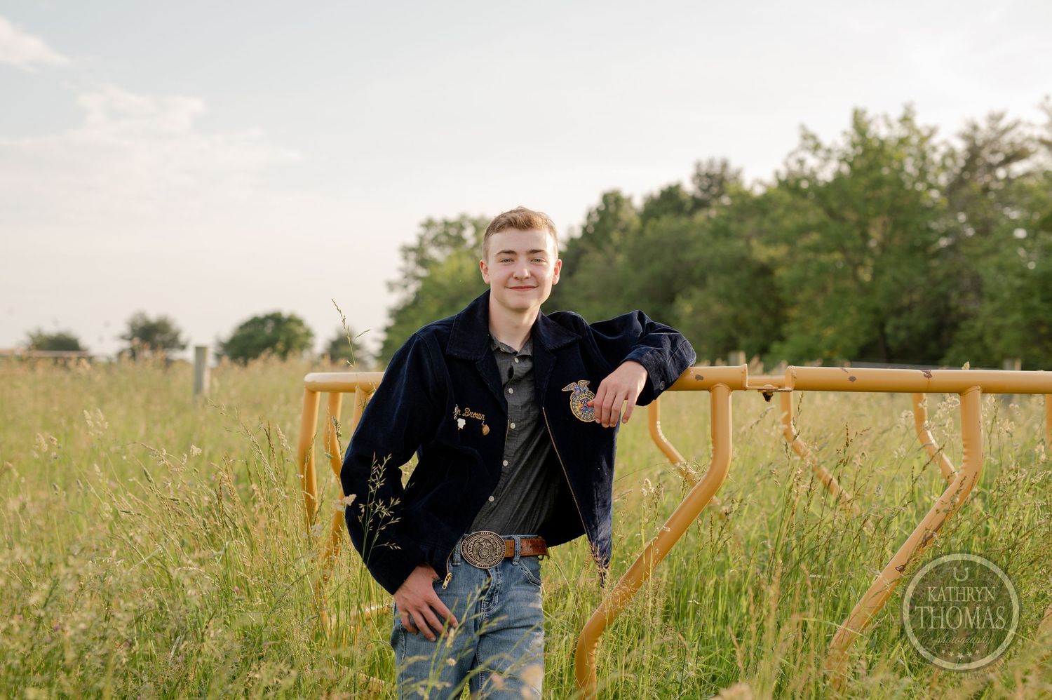 Individual in dark jacket leans casually against yellow railing in summer meadow.