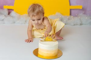 you are my sunshine cake smash session. toddler girl wearing yellow dress sitting in front of pink and yellow background