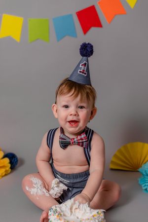 first birthday cake smash with boy sitting in front of grey and colourful background. boy is wearing a party hat