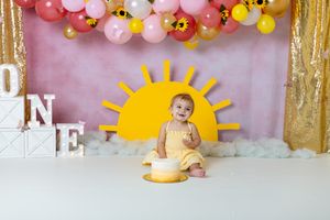 you are my sunshine cake smash session. toddler girl wearing yellow dress sitting in front of pink and yellow background