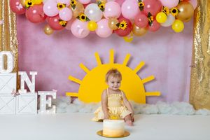 you are my sunshine cake smash session. toddler girl wearing yellow dress sitting in front of pink and yellow background