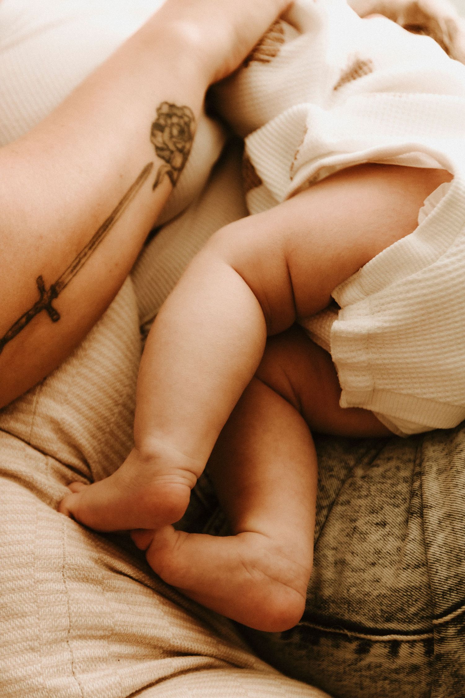 Close-up view of baby's bare feet resting on soft bedding next to arm with rose and cross tattoos.