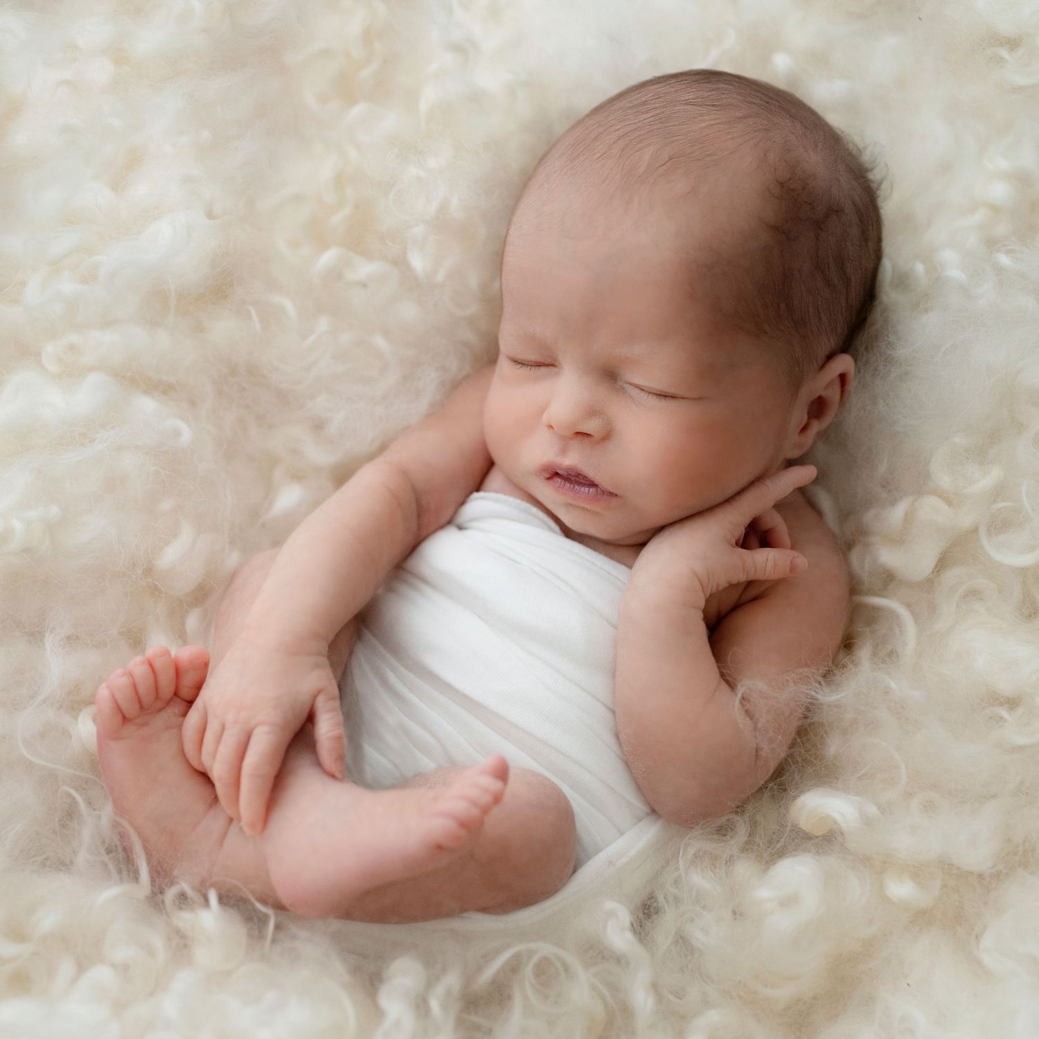 A peaceful newborn baby sleeping soundly on soft white fur blanket in natural light.