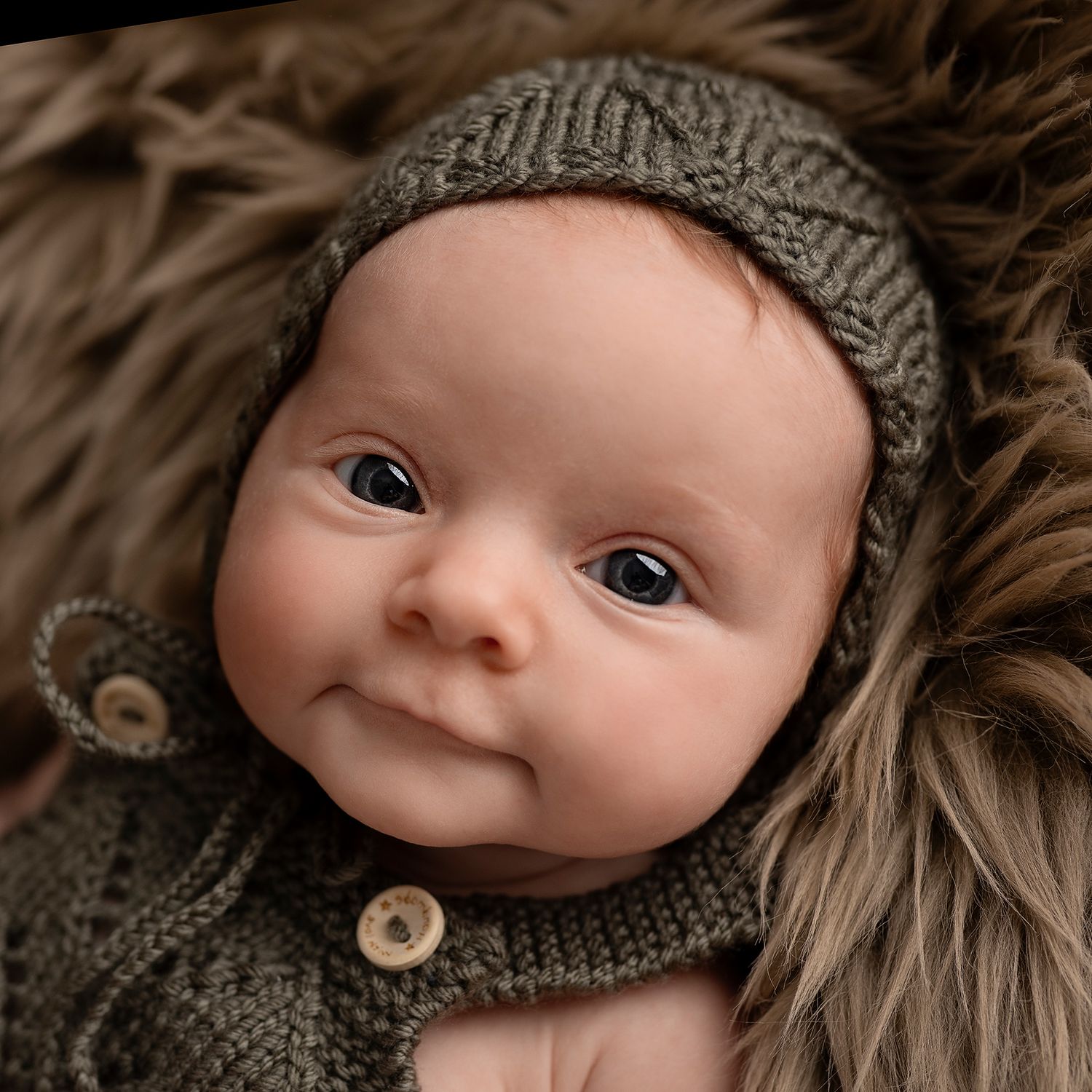 Sweet baby making eye contact and wearing a green knit hat and romper lying on soft brown fur blanket.