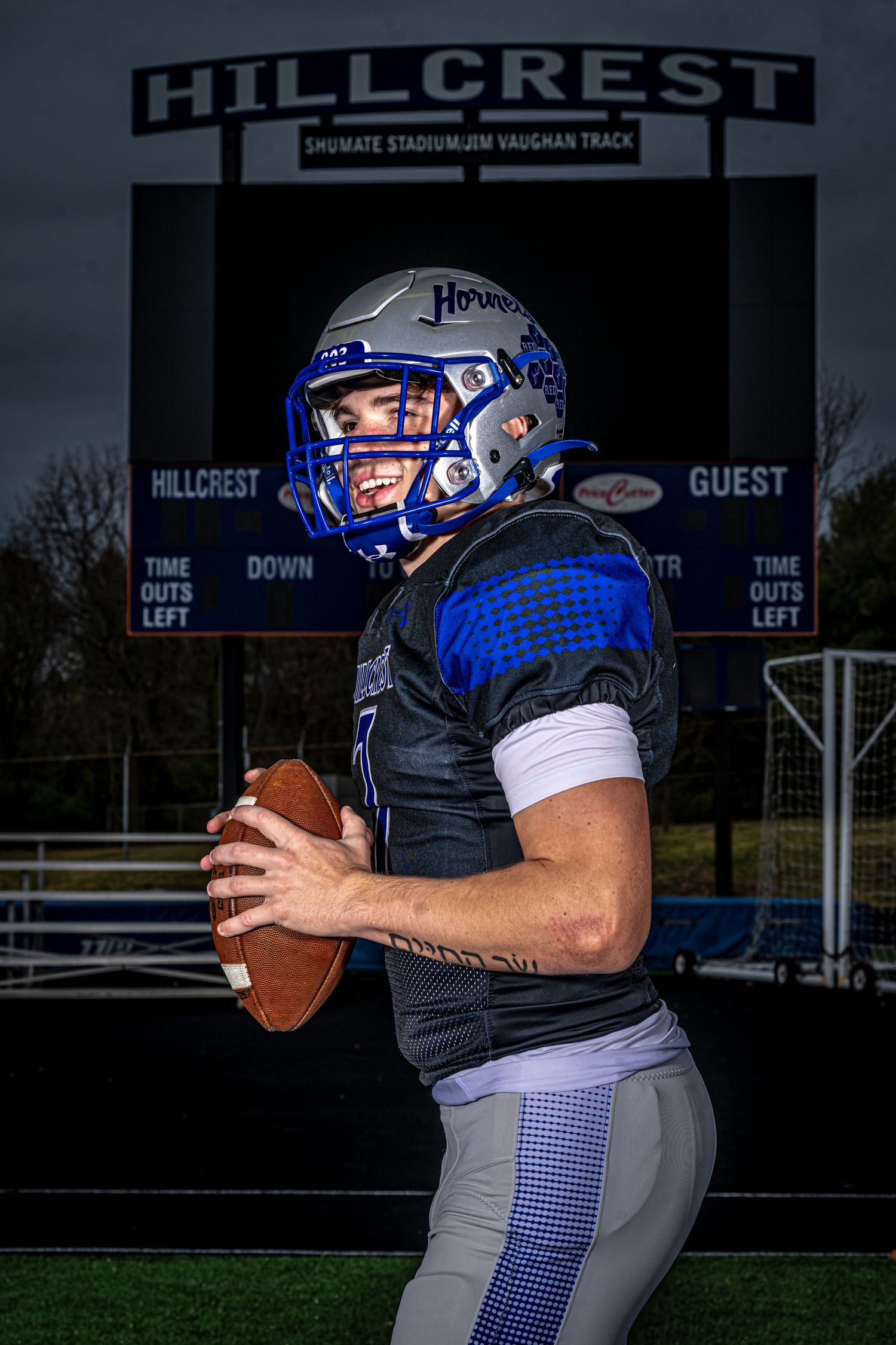 Football player in blue and grey uniform holds football on field with Hillcrest stadium sign visible in background.