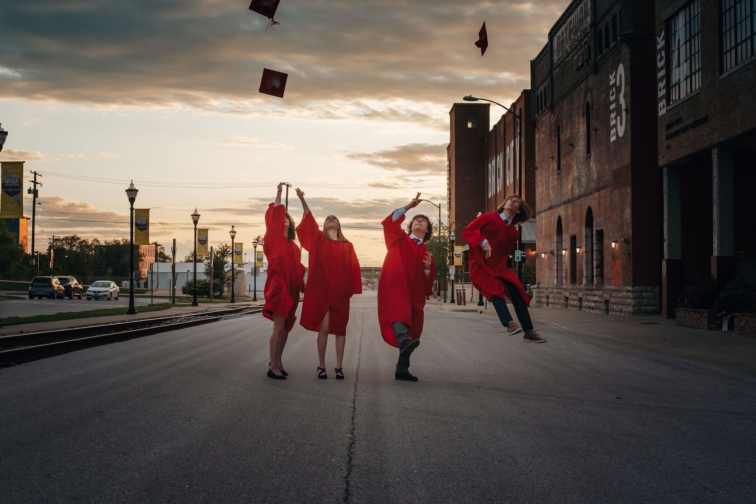 Graduates in red gowns throwing caps in the air at sunset against urban backdrop.